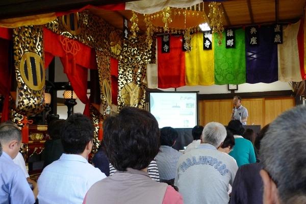 赤、黄、緑、青などののれんがかかる室内に、たくさんの人々が座って、マイクを持つ男性の話を聞いている写真